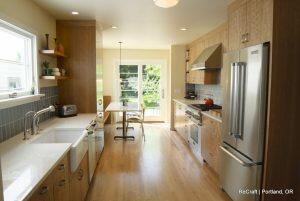 A modern kitchen with wooden cabinets, white countertops, stainless steel appliances, a double sink, and a dining area near a glass door leading to an outdoor space.