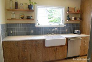 A modern kitchen features wooden cabinets, a white farmhouse sink, a dishwasher, and open shelves with various jars and plants. A window above the sink provides natural light.
