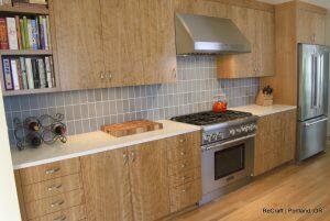 Modern kitchen with wooden cabinets, stainless steel appliances, tiled backsplash, and a countertop featuring a chopping board and a knife block. A wine rack with bottles is placed on the counter.