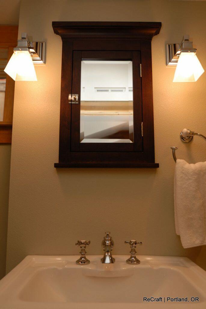 A bathroom vanity with a dark wood-framed mirror, two wall-mounted lights, a white sink with silver faucet handles, and a towel ring holding a white towel. Text reads: "ReCraft | Portland, OR. A bathroom vanity with a dark wood-framed mirror, two wall-mounted lights, a white sink with silver faucet handles, and a towel ring holding a white towel. Text reads: "ReCraft | Portland, OR.