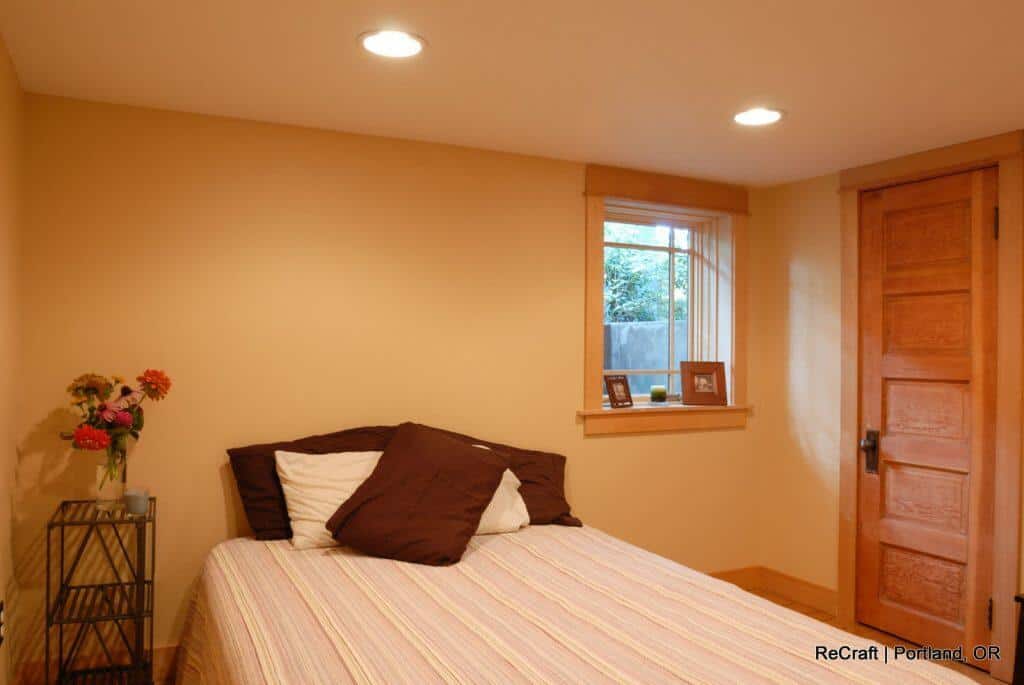 A small bedroom with beige walls features a bed with striped bedding, brown and white pillows, a wooden door, a window with a view of greenery, and a side table with flowers in a vase. A small bedroom with beige walls features a bed with striped bedding, brown and white pillows, a wooden door, a window with a view of greenery, and a side table with flowers in a vase.
