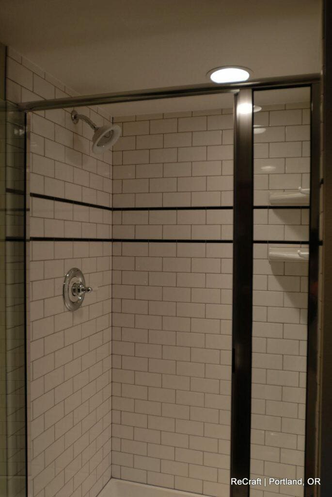 A tiled shower with white subway tiles, a glass door, chrome fixtures, and a recessed light above. The tiles have a black border in the middle. Sign reads "ReCraft | Portland, OR". A tiled shower with white subway tiles, a glass door, chrome fixtures, and a recessed light above. The tiles have a black border in the middle. Sign reads "ReCraft | Portland, OR".