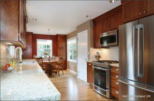 A modern kitchen with stainless steel appliances, wooden cabinets, granite countertops, and a dining area with a table and chairs near a window.