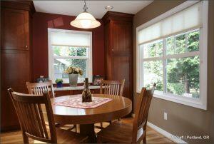 A dining area with a round wooden table and four chairs, against a backdrop of red and brown walls, a window with a view of greenery, and wall cabinets. A pendant light hangs above the table.
