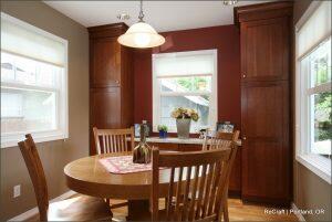 A dining area with a round wooden table and four chairs, set against a red and beige wall with large cabinets, windows, and vase of flowers. A hanging light fixture is positioned above the table.