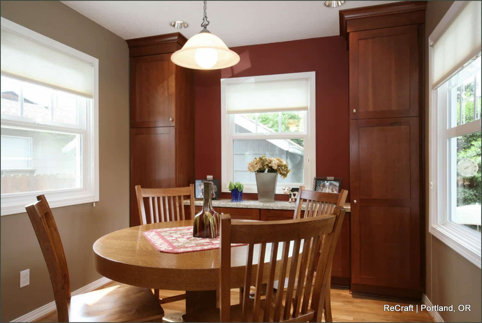A dining area with a round wooden table and four chairs, set against a red and beige wall with large cabinets, windows, and vase of flowers. A hanging light fixture is positioned above the table. A dining area with a round wooden table and four chairs, set against a red and beige wall with large cabinets, windows, and vase of flowers. A hanging light fixture is positioned above the table.