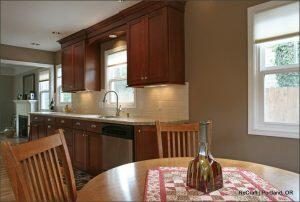 A kitchen with wooden cabinets, a double sink, white tile backsplash, and a window above the sink. A wooden dining table with a decorative bottle and placemat is in the foreground.