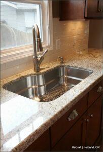 A modern kitchen sink with a stainless steel faucet set in a granite countertop. The backsplash is white tile. Cabinets are dark wood. A window above provides natural light.
