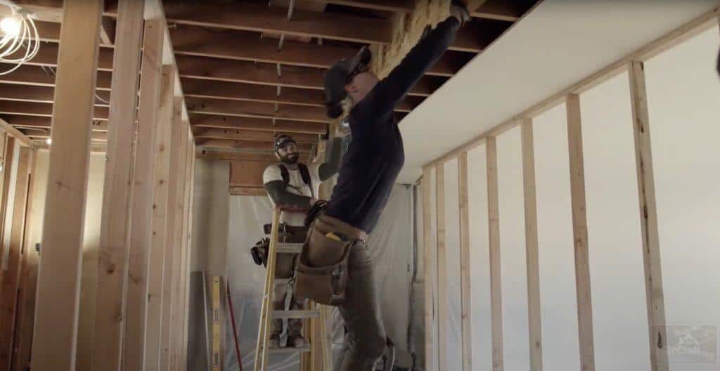 Two construction workers install insulation and drywall in a building with exposed wooden framing. One worker stands on a ladder while the other reaches overhead. Tools and materials are scattered nearby. Two construction workers install insulation and drywall in a building with exposed wooden framing. One worker stands on a ladder while the other reaches overhead. Tools and materials are scattered nearby.