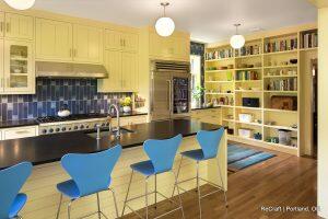 Modern kitchen with yellow cabinets, a large black countertop island with seating, stainless steel appliances, blue backsplash tiles, open shelves with books, and three blue chairs. This Irvington kitchen remodel perfectly blends style and functionality.