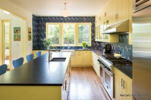 A modern Irvington kitchen remodel features yellow cabinets, black countertops, and stainless steel appliances. Blue wall tiles add a vibrant touch, while a central island with a sink enhances functionality. In the background, a hallway and artworks complete the look.