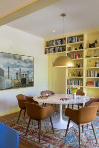 A modern dining area with a round white table, four brown chairs, a hanging light fixture, a bookshelf filled with books and decor, and a large painting of an industrial scene.