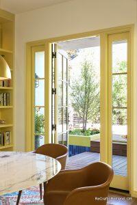 Open yellow-framed French doors lead from a cozy interior space with a marble table to a sunny outdoor patio with greenery and colorful planters.