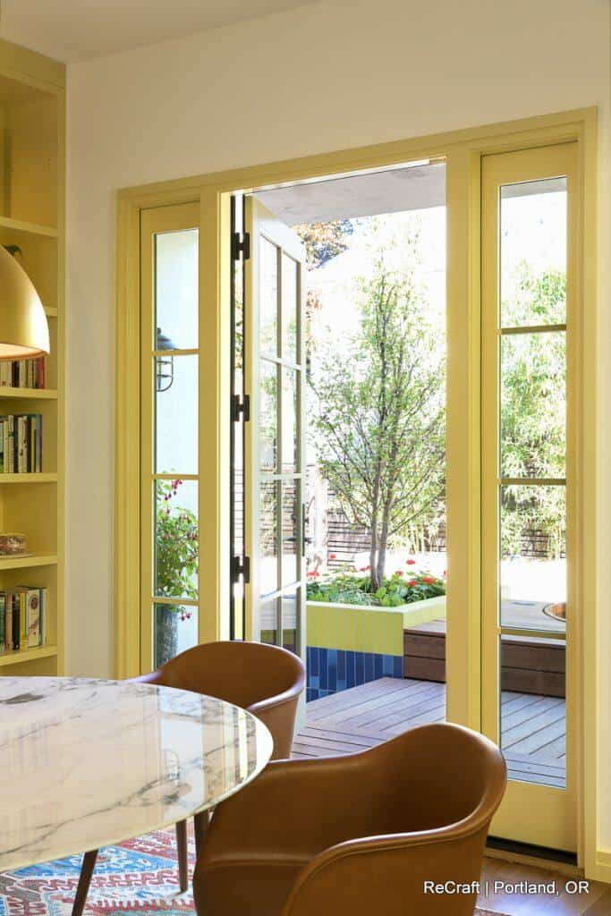 Open yellow-framed French doors lead from a cozy interior space with a marble table to a sunny outdoor patio with greenery and colorful planters. Open yellow-framed French doors lead from a cozy interior space with a marble table to a sunny outdoor patio with greenery and colorful planters.