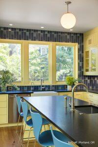 A modern kitchen with yellow cabinets, a large black countertop island, blue barstools, and a backsplash of vertical blue tiles. A round pendant light hangs above the island.