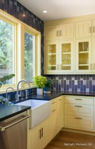 A modern kitchen with yellow cabinets, black countertops, blue tiled backsplash, farmhouse sink, large windows, and potted plants. Kitchen designed by ReCraft in Portland, OR.