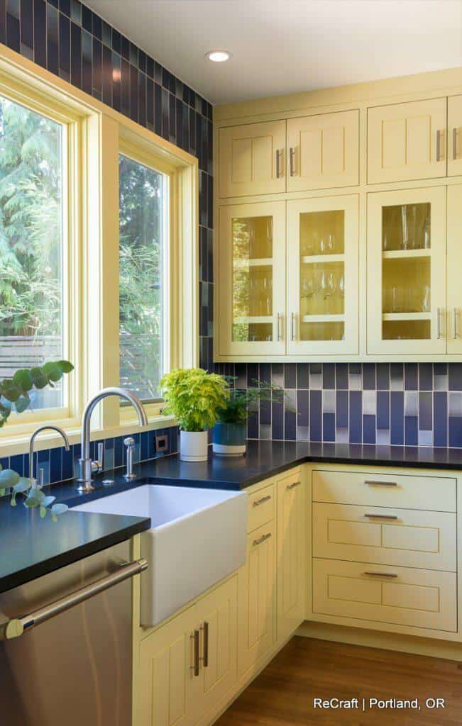 A modern kitchen with yellow cabinets, black countertops, blue tiled backsplash, farmhouse sink, large windows, and potted plants. Kitchen designed by ReCraft in Portland, OR. A modern kitchen with yellow cabinets, black countertops, blue tiled backsplash, farmhouse sink, large windows, and potted plants. Kitchen designed by ReCraft in Portland, OR.