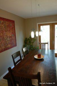 A wooden dining table with six chairs is centered in a room. A red tapestry hangs on the wall, three pendant lights hang above the table, and a glass door leads outside. A plant is in the corner.