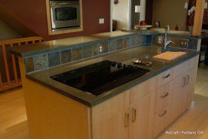 A modern kitchen with wood cabinets, a stovetop on an island, a built-in oven, and a grey countertop with a tiled backsplash. The image is credited to ReCraft in Portland, OR.