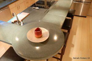 A modern kitchen features a round greenish-gray countertop extension with a small dish containing a red item, next to a sink and stove. Dark wooden bar stools are placed in front of the counter.