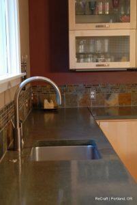A modern kitchen countertop with a stainless steel faucet and sink. There is a backsplash with brown, green, and tan tiles, and a partially open cabinet displaying glassware in the background.