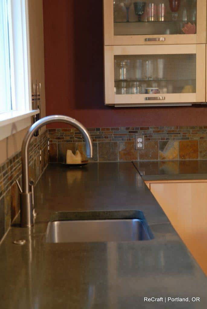 A modern kitchen countertop with a stainless steel faucet and sink. There is a backsplash with brown, green, and tan tiles, and a partially open cabinet displaying glassware in the background. A modern kitchen countertop with a stainless steel faucet and sink. There is a backsplash with brown, green, and tan tiles, and a partially open cabinet displaying glassware in the background.