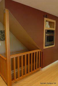 A kitchen with an oven built into a wall niche, located under a sloped ceiling next to a wooden railing. Books are stored above the oven. The setting is ReCraft in Portland, OR.