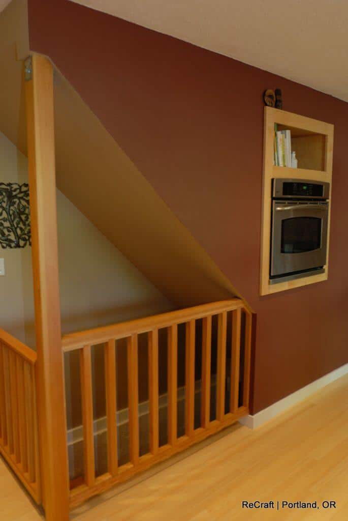 A kitchen with an oven built into a wall niche, located under a sloped ceiling next to a wooden railing. Books are stored above the oven. The setting is ReCraft in Portland, OR. A kitchen with an oven built into a wall niche, located under a sloped ceiling next to a wooden railing. Books are stored above the oven. The setting is ReCraft in Portland, OR.