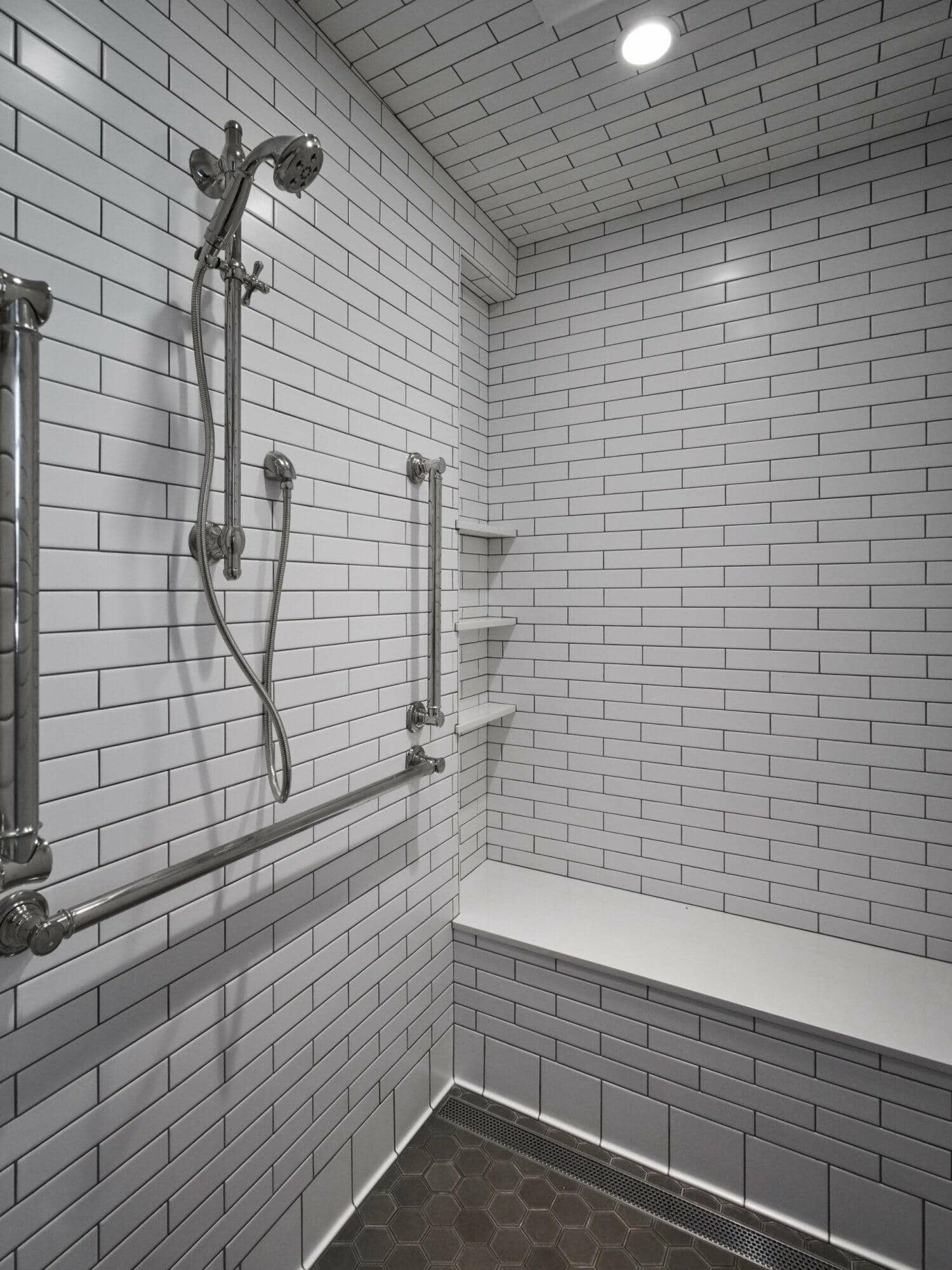 A modern shower with white subway tiles, a handheld showerhead, grab bars, a bench, and built-in corner shelves.