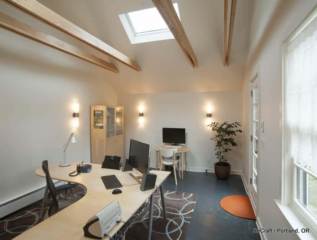 A modern office space with a skylight, multiple desks, computers, a potted plant, and wooden ceiling beams, illuminated by wall-mounted lights. A modern office space with a skylight, multiple desks, computers, a potted plant, and wooden ceiling beams, illuminated by wall-mounted lights.
