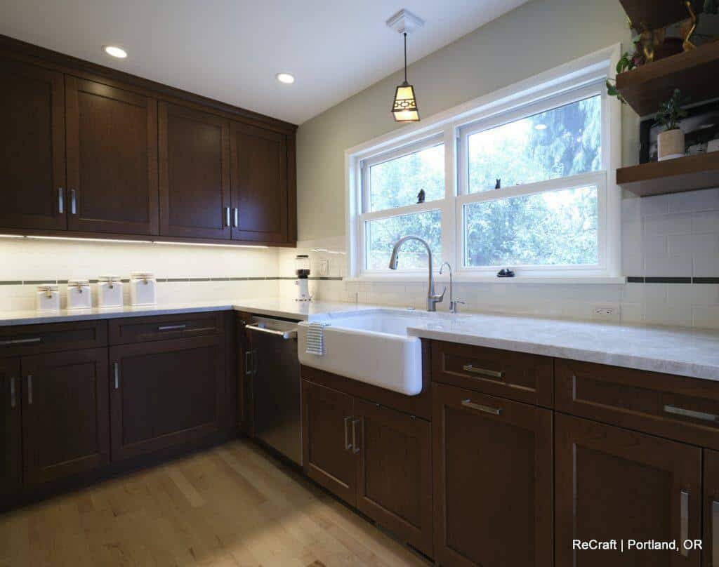 A modern kitchen features dark wooden cabinets, light countertops, a farmhouse sink, stainless steel dishwasher, and large windows. Shelves on the right hold various items. Light wood floor contrasts the cabinetry. A modern kitchen features dark wooden cabinets, light countertops, a farmhouse sink, stainless steel dishwasher, and large windows. Shelves on the right hold various items. Light wood floor contrasts the cabinetry.