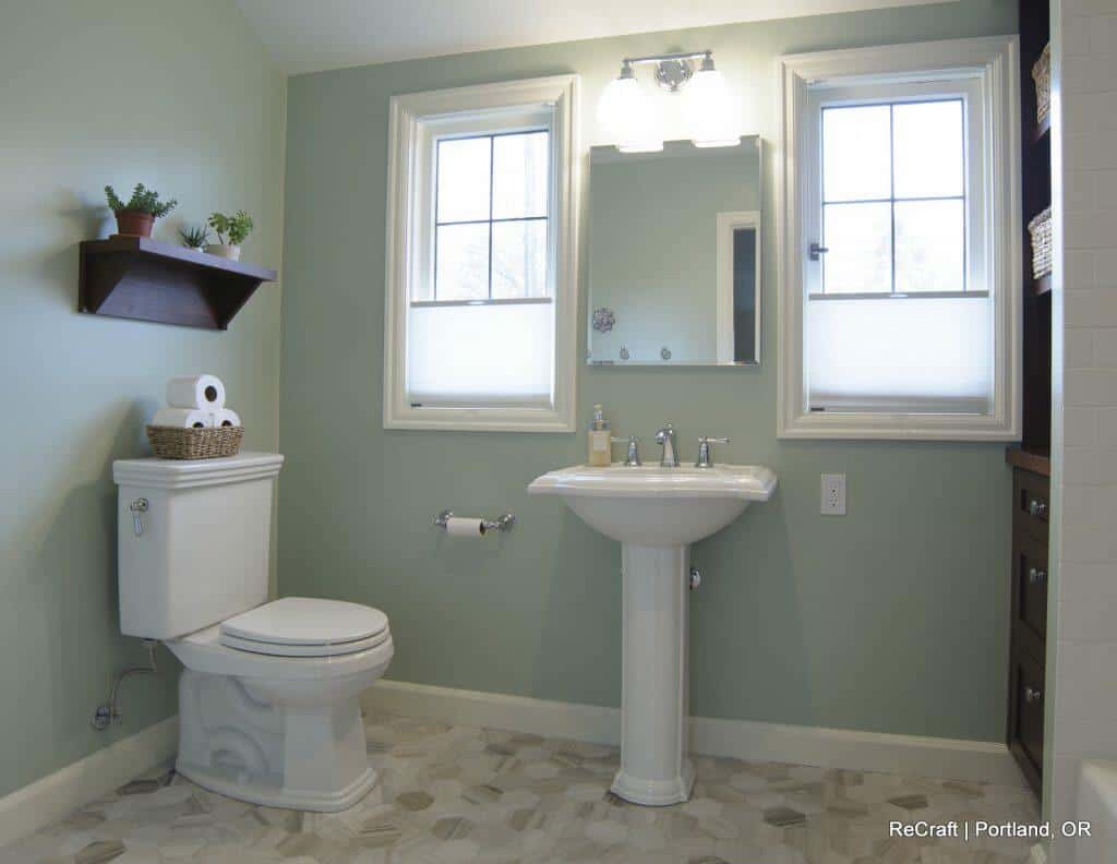 A modern bathroom with green walls, featuring a white toilet, pedestal sink, wall-mounted shelf, and twin windows above the sink. A mirrored cabinet is adjacent. Floor tiles are patterned. Text reads "ReCraft | Portland, OR". A modern bathroom with green walls, featuring a white toilet, pedestal sink, wall-mounted shelf, and twin windows above the sink. A mirrored cabinet is adjacent. Floor tiles are patterned. Text reads "ReCraft | Portland, OR".