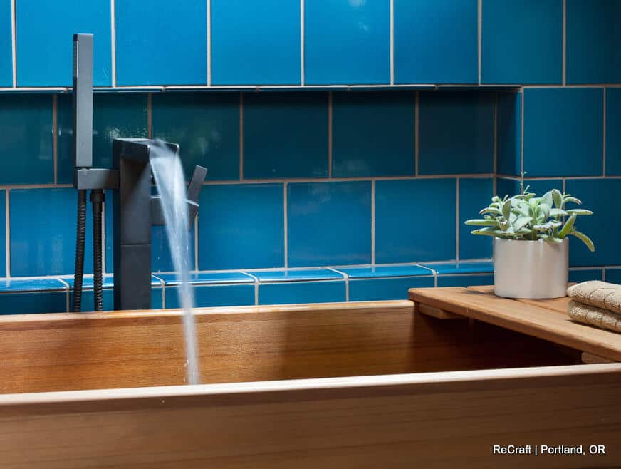 A wooden bathtub with water pouring from a modern faucet against a blue tiled wall. A potted plant and folded towels sit on a wooden ledge. Text at the bottom right: "ReCraft | Portland, OR. A wooden bathtub with water pouring from a modern faucet against a blue tiled wall. A potted plant and folded towels sit on a wooden ledge. Text at the bottom right: "ReCraft | Portland, OR.