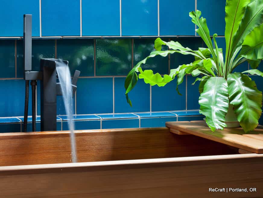 A wooden bathtub with running water, positioned against a blue tile wall with a green plant on a wooden shelf. A wooden bathtub with running water, positioned against a blue tile wall with a green plant on a wooden shelf.