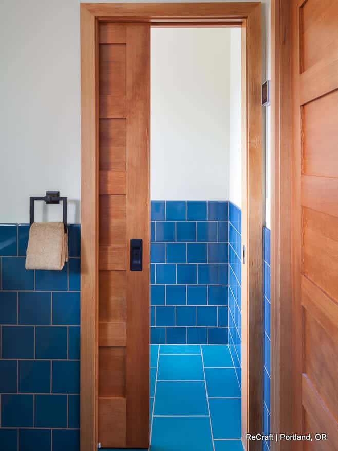 A bathroom with blue tiled walls and floor features a sliding wooden door. A dark towel rack with a beige towel is mounted on the left wall. A bathroom with blue tiled walls and floor features a sliding wooden door. A dark towel rack with a beige towel is mounted on the left wall.