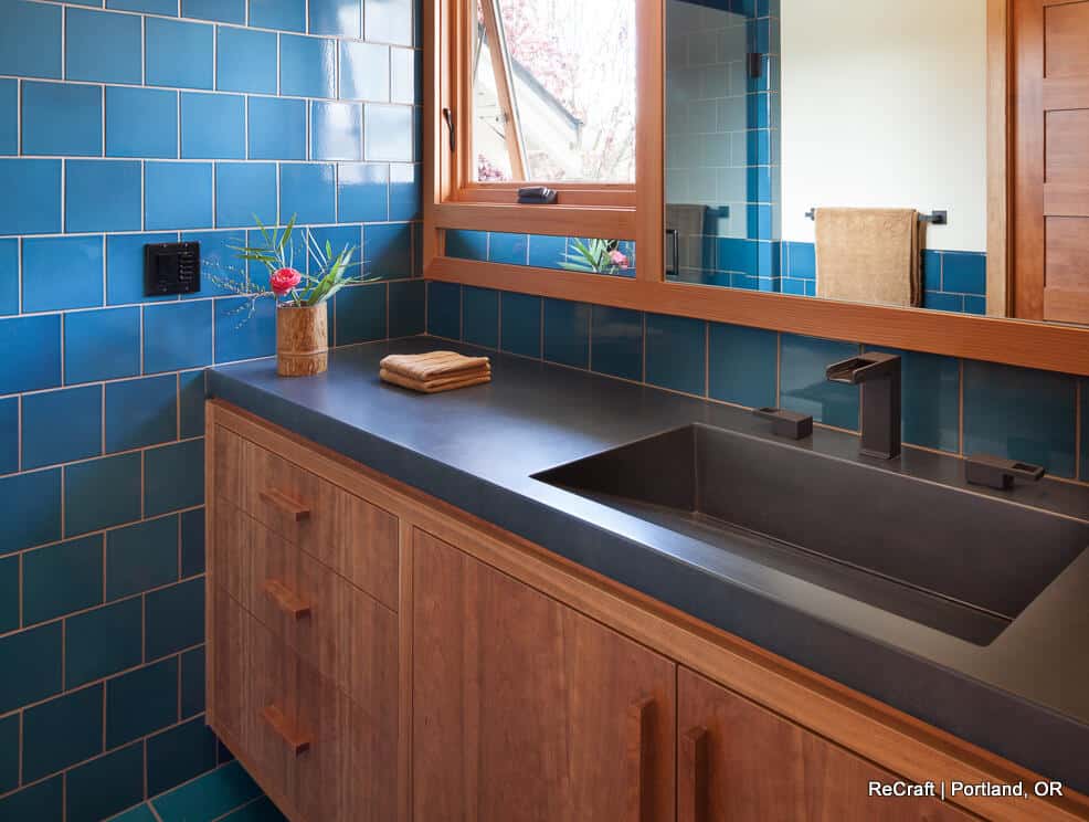 A bathroom with blue tiled walls, a black countertop, a wooden cabinet with drawers, a rectangular sink, a mirror, and a window. A small plant and folded hand towels are on the counter. A bathroom with blue tiled walls, a black countertop, a wooden cabinet with drawers, a rectangular sink, a mirror, and a window. A small plant and folded hand towels are on the counter.
