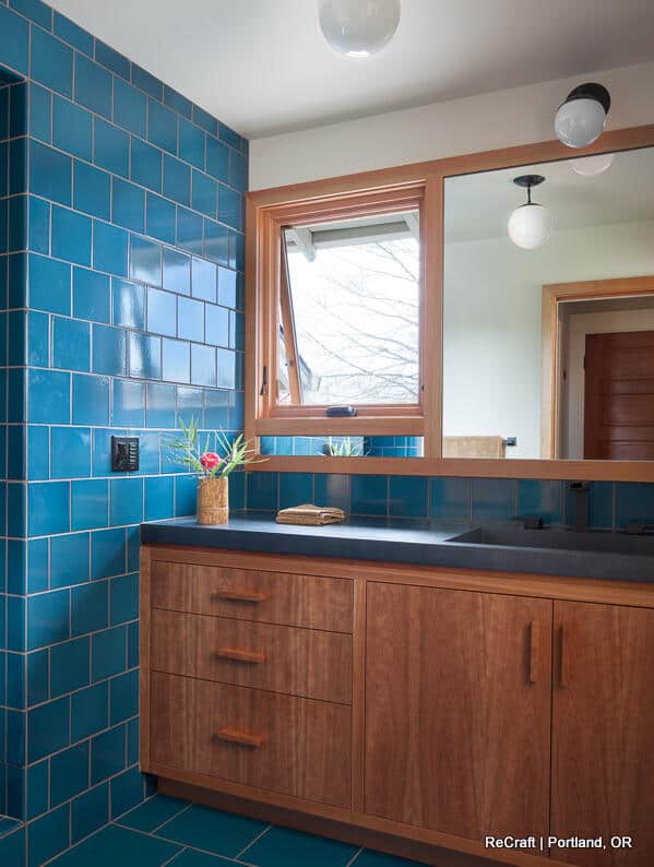 A bathroom with blue tiled walls, wooden cabinetry, a black countertop with a towel, a plant in a vase, a large mirror, and two spherical lights hanging from the ceiling. A bathroom with blue tiled walls, wooden cabinetry, a black countertop with a towel, a plant in a vase, a large mirror, and two spherical lights hanging from the ceiling.