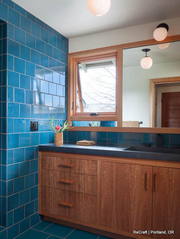A bathroom with blue tiled walls, wooden vanity with dark countertop, a large mirror, and modern light fixtures. A bathroom with blue tiled walls, wooden vanity with dark countertop, a large mirror, and modern light fixtures.