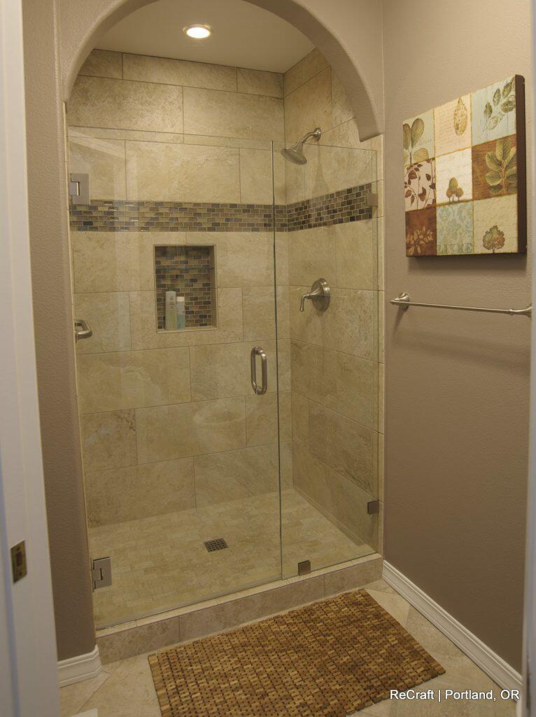 A modern shower with tiled walls, a built-in niche with products, glass door, and overhead lighting. A towel rack is on the wall beside it, with a small rug on the floor. Artwork hangs on the adjacent wall. A modern shower with tiled walls, a built-in niche with products, glass door, and overhead lighting. A towel rack is on the wall beside it, with a small rug on the floor. Artwork hangs on the adjacent wall.