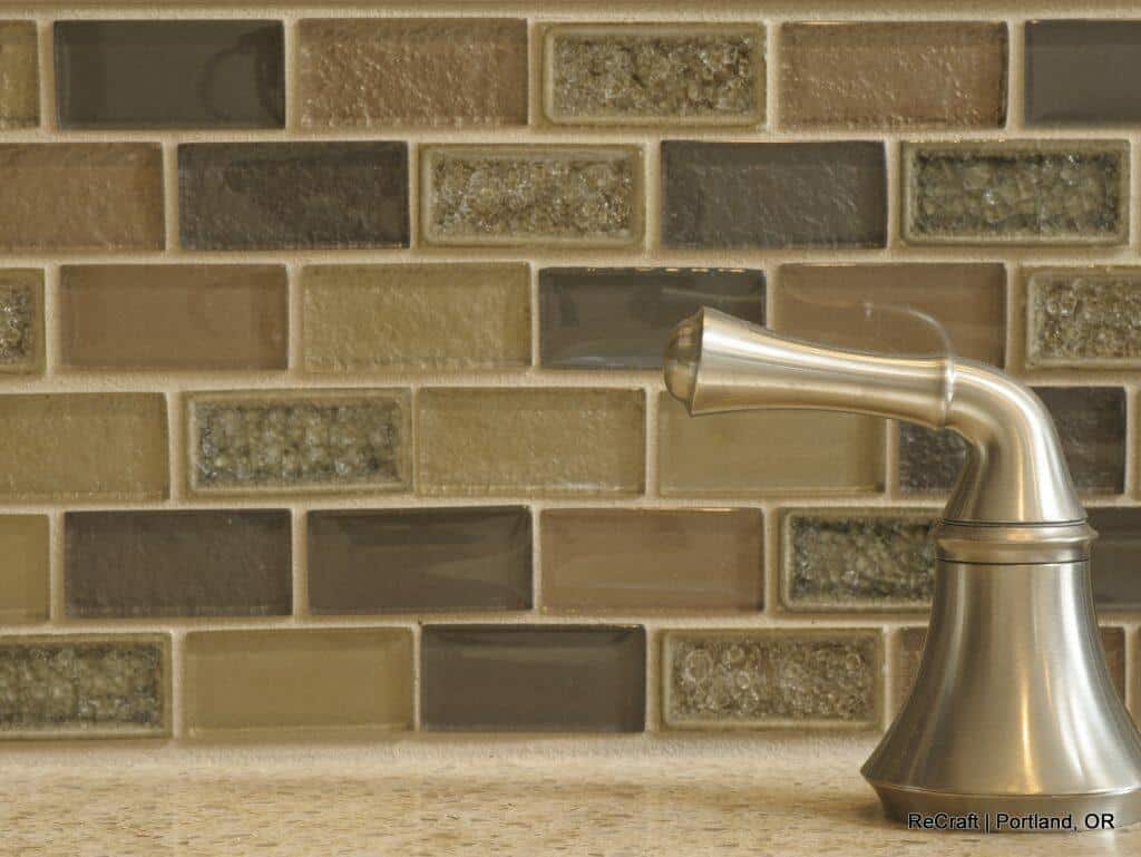 Close-up of a stainless steel kitchen faucet with a beige granite countertop and a mosaic tile backsplash in shades of brown, gray, and beige. Close-up of a stainless steel kitchen faucet with a beige granite countertop and a mosaic tile backsplash in shades of brown, gray, and beige.