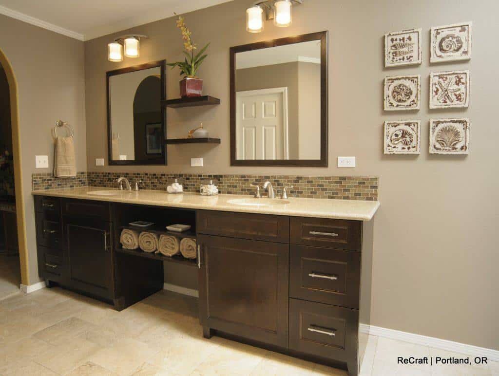 A modern bathroom with a double sink vanity, dark wood cabinets, wall-mounted shelves, and two mirrors. The beige-tiled floor and backsplash contrast with the neutral wall color. ReCraft | Portland, OR. A modern bathroom with a double sink vanity, dark wood cabinets, wall-mounted shelves, and two mirrors. The beige-tiled floor and backsplash contrast with the neutral wall color. ReCraft | Portland, OR.