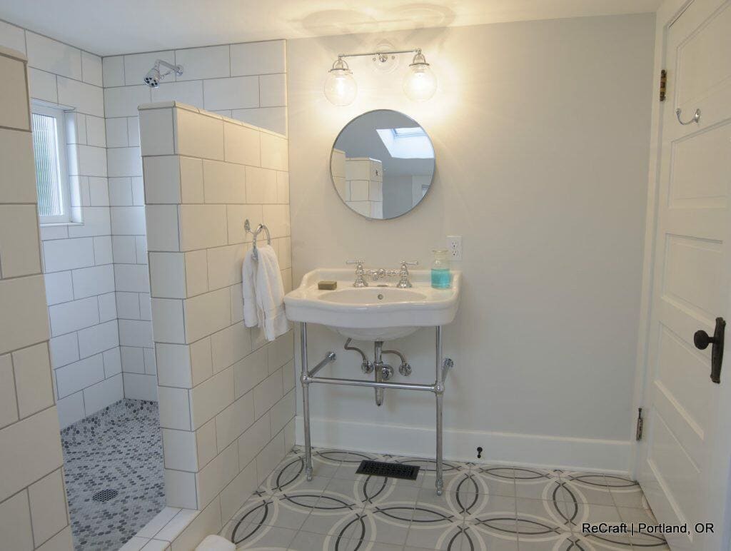 A modern bathroom with a white sink, round mirror, glass bottles, towel, and walk-in shower with a tiled floor. The walls and floor feature white tiles, and the room is well-lit with two light fixtures. A modern bathroom with a white sink, round mirror, glass bottles, towel, and walk-in shower with a tiled floor. The walls and floor feature white tiles, and the room is well-lit with two light fixtures.