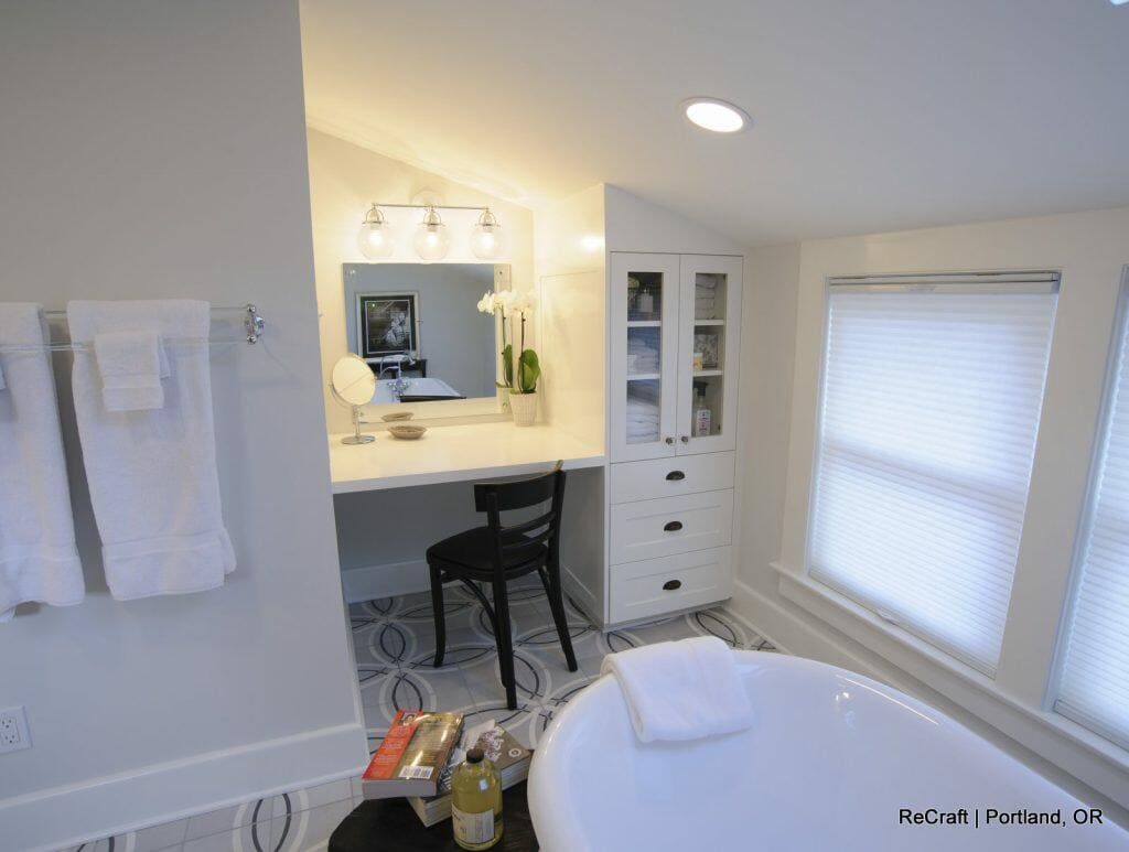 A modern bathroom with a white vanity and chair, backlit mirror, cabinets, towel rack with two towels, and a freestanding bathtub with a tray holding a book and a bottle. A modern bathroom with a white vanity and chair, backlit mirror, cabinets, towel rack with two towels, and a freestanding bathtub with a tray holding a book and a bottle.
