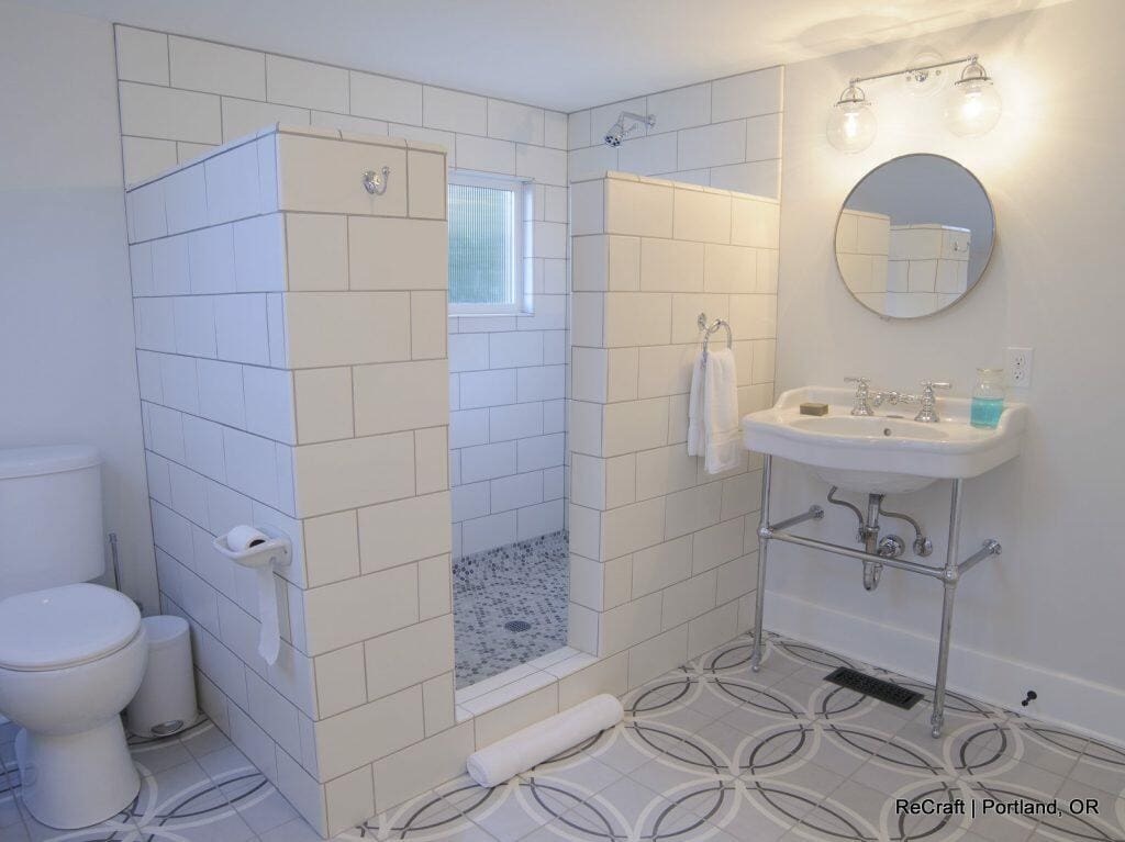 A bathroom with a walk-in shower featuring white tiled walls, a toilet, a sink with a round mirror above it, a towel hanging, and patterned floor tiles. A small window is inside the shower area. A bathroom with a walk-in shower featuring white tiled walls, a toilet, a sink with a round mirror above it, a towel hanging, and patterned floor tiles. A small window is inside the shower area.