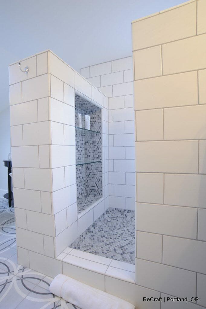 A white-tiled shower with built-in shelves containing toiletries. The floor and wall tiles have a mix of hexagonal patterns. A towel is folded on the floor outside the shower. Text in the corner reads "ReCraft | Portland, OR. A white-tiled shower with built-in shelves containing toiletries. The floor and wall tiles have a mix of hexagonal patterns. A towel is folded on the floor outside the shower. Text in the corner reads "ReCraft | Portland, OR.