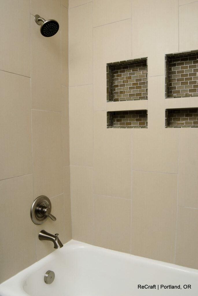 Shower area with beige tiles, a black showerhead, and dual square shelves with dark mosaic tile backing. Chrome faucet and handle above white bathtub. Text: "ReCraft | Portland, OR" at bottom right. Shower area with beige tiles, a black showerhead, and dual square shelves with dark mosaic tile backing. Chrome faucet and handle above white bathtub. Text: "ReCraft | Portland, OR" at bottom right.