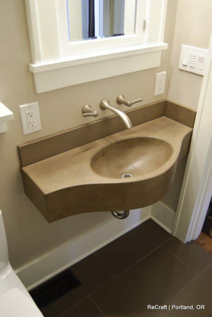 A bathroom sink with a wall-mounted faucet and a unique curved countertop design. The vanity is next to a white toilet, and a mirror is positioned above the sink. Electrical outlets are nearby. A bathroom sink with a wall-mounted faucet and a unique curved countertop design. The vanity is next to a white toilet, and a mirror is positioned above the sink. Electrical outlets are nearby.