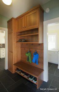 A wooden mudroom with storage cabinets, open shelves, and hooks holding jackets. A bench with shoe cubbies underneath displays various shoes. A doorway leads to a kitchen area in the background.