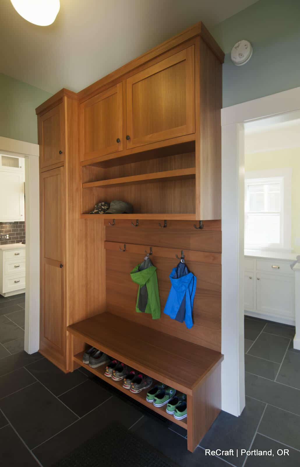 A wooden mudroom with storage cabinets, open shelves, and hooks holding jackets. A bench with shoe cubbies underneath displays various shoes. A doorway leads to a kitchen area in the background. A wooden mudroom with storage cabinets, open shelves, and hooks holding jackets. A bench with shoe cubbies underneath displays various shoes. A doorway leads to a kitchen area in the background.