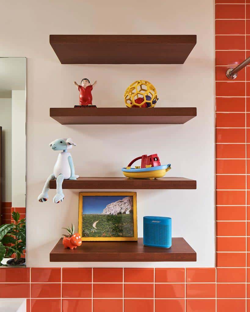 Three wooden shelves holding a red figurine, a yellow ball, a blue stuffed animal, a toy boat, a framed photo, a small orange figure, and a blue speaker against an orange-tiled wall. Three wooden shelves holding a red figurine, a yellow ball, a blue stuffed animal, a toy boat, a framed photo, a small orange figure, and a blue speaker against an orange-tiled wall.